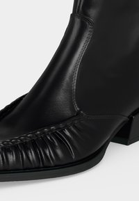 Bottine noire en cuir avec un détail froncé sur l'orteil, des panneaux cousus et un petit talon bloc sur fond blanc.