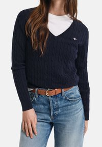 Pull en maille torsadée bleu marine avec un col en V, porté sur un t-shirt blanc. Associé à un jean bleu clair et une ceinture marron avec une boucle argentée.