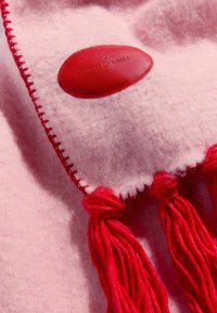 Różowy wełniany koc z czerwoną skórzaną metką, kontrastującym czerwonym ściegiem i frędzlami z czerwonymi pomponami. Miękka faktura i ciepły wygląd.
