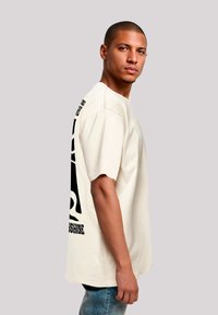 Light beige oversized t-shirt featuring bold black graphic designs on the back, with short sleeves and a relaxed fit. Denim pants worn.