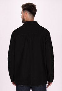 Veste noire, en tissu lisse, avec un col et des manches longues. La vue de dos montre un design simple sans motifs ou accents supplémentaires.