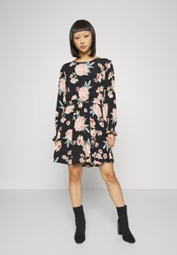 Vestido preto de manga longa com padrão floral em rosa e verde, cintura ajustada e saia em camadas. Combinado com botas pretas até ao tornozelo.