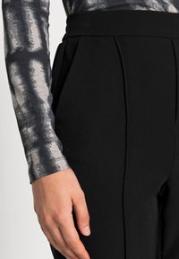 Calças pretas feitas à medida com pregas marcadas, combinadas com uma blusa cinza de mangas compridas com um padrão abstrato metálico. Close-up na posição das mãos.
