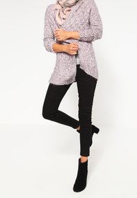 Ljust lila, fluffig cardigan med långa ärmar, öppen front och sidoflikar, kombinerad med svarta tajta byxor och svarta ankelskor.