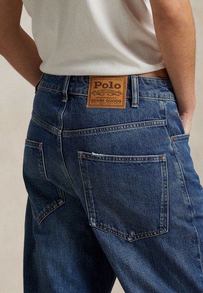 Persona que lleva puestos unos vaqueros vaqueros azul Polo Ralph Lauren con una etiqueta de cuero visible en la cintura trasera, camisa blanca por dentro.