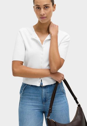 Frau trägt ein weißes, kurzärmeliges, mit Knöpfen versehenes Oberteil und blaue Jeans, hält eine dunkelbraune Lederhandtasche an ihrem Henkel.