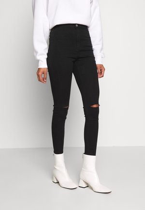 Black skinny jeans with high waist, featuring knee rips. Paired with white ankle boots, smooth texture, and a fitted silhouette.