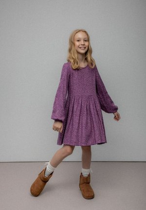 Fioletowa sukienka w kwiaty z długimi rękawami i marszczonym dołem; do niej brązowe botki za kostkę i białe skarpetki. Gładkie tło.