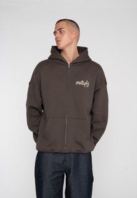 Brun lynlås hoodie med stor hætte og forlommer. Har et diskret grafisk logo på venstre brystområde. Blød stoftekstur.