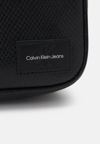 Borsa nera testurizzata con design in pelle di serpente. Presenta un'etichetta con il logo Calvin Klein Jeans in bianco su sfondo nero.