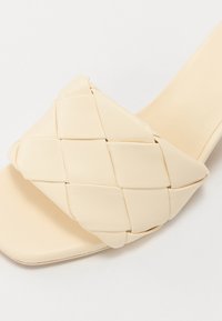 Beige leather slide sandal with a woven upper design, featuring a square toe and smooth, matte texture.