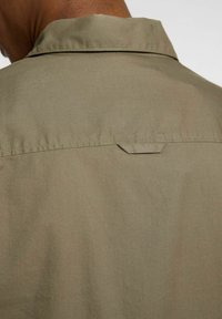 Camicetta verde oliva con una texture liscia, caratterizzata da un'apertura posteriore e un piccolo bottone; design minimalista senza motivi o accenti visibili.