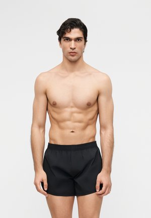 Postaven mlad moški s temnimi kodrastimi lasmi stoji obrnjen proti kameri, brez majice, oblečene v navadne črne kopalne hlače na enostavni svetli podlagi.