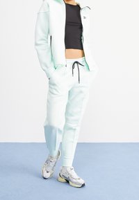 Light mint green athletic tracksuit with a zip-up jacket and drawstring pants. Black cropped top underneath. Gray and silver sneakers.