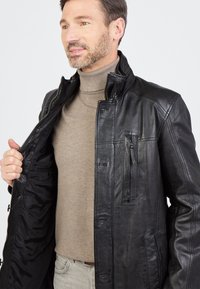 Schwarze Lederjacke mit einem Reißverschluss vorne, stehendem Kragen und mehreren Taschen. Getragen über einem beigen Rollkragenpullover, der eine glatte Textur zeigt.