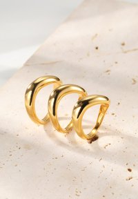 Trzy złote pierścionki o gładkim, zaokrąglonym wzornictwie, z wypolerowanym wykończeniem. Są wyeksponowane na teksturowanej, jasnej powierzchni kamienia.