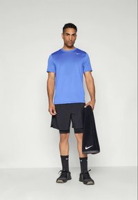 Mand i blå athletisk skjorte, sorte shorts og sorte sneakers står og holder et sort Nike håndklæde mod en ensfarvet baggrund.