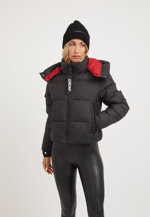 Mujer con chaqueta acolchada negra con forro rojo, gorro negro y leggings negros brillantes, de pie con una mano en el bolsillo.