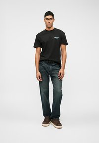 Juodas medvilninis marškinėliai su priekiu iliustracija, mėlyna spalva; derinami su tamsiai mėlynomis džinsomis ir rudos spalvos raišteliu suvertais sportbačiais. Modelis stovi priešais baltą foną.