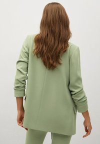 Vaaleanvihreä räätälöity blazer, jossa on rento mitoitus, käärityt hihat ja takaventti. Valmistettu sileästä kankaasta ilman näkyviä kuvioita.