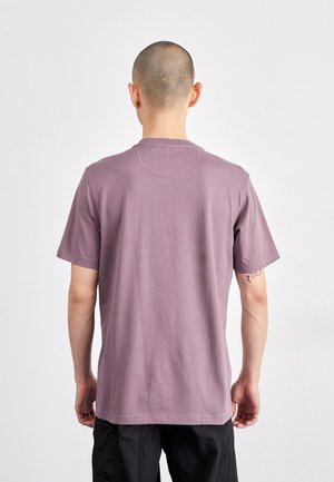 Mann mit rasiertem Kopf, der wegschaut, trägt ein schlichtes kurzärmeliges T-Shirt in Mauve und schwarze Hose, steht vor weißem Hintergrund.