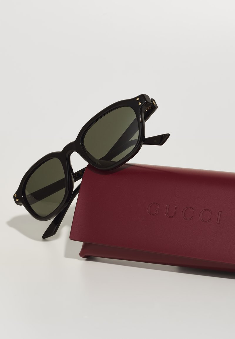Lunettes de soleil noires surdimensionnées avec des verres sombres et des accents dorés, posées sur un étui Gucci bordeaux, comportant un logo en relief.