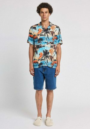 Homme debout portant une chemise à manches courtes à imprimé tropical coloré, un short en jean bleu et des chaussures beige sans lacets sur un fond uni.