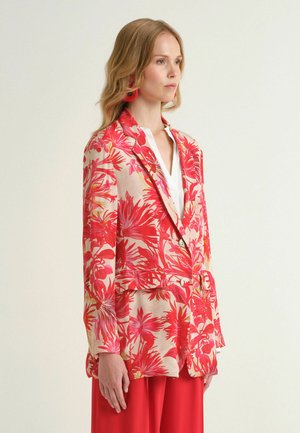 Femme portant un chemisier blanc sous un blazer floral rouge et beige avec un pantalon taille haute assorti, debout de profil devant un fond uni.