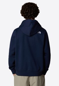 Sudadera con capucha de color azul marino, fabricada en tejido suave, con puños y dobladillo acanalados, y un pequeño logo blanco en el hombro trasero.
