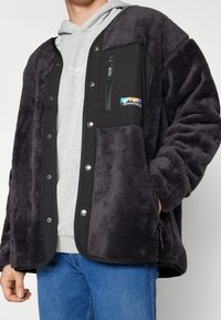 Uomo che indossa una giacca in fleece grigio scuro con bottoni a pressione sopra un hoodie grigio chiaro e jeans blu, con una mano nella tasca della giacca.