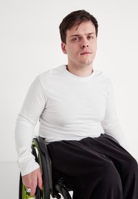 Fehér hosszúujjú ing, fekete nadrág, kerekesszékben ül. A kerekesszék fekete kerekekkel és zöld részletekkel rendelkezik. Simogatós anyag textúra.