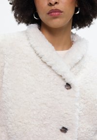 Κρεμ παλτό από δερμάτινη γούνα με γιακά με κοψίματα, διαθέτοντας δύο καφέ κουμπιά και μαλακή, υφασμάτινη επιφάνεια.