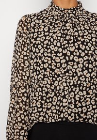Schwarze Bluse mit einem ganzflächigen beigen und braunen Leopardenmuster. Verfügt über einen gefalteten hohen Kragen und lange, eng anliegende Ärmel. Leichter Stoff.