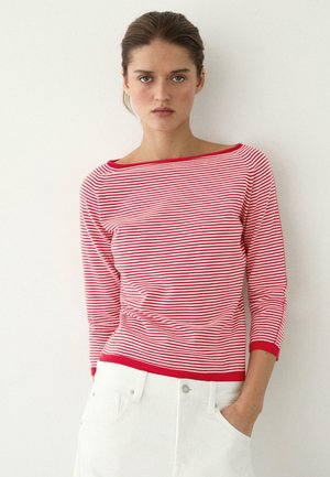 Donna con capelli castani, indossa una maglia a maniche lunghe a righe rosse e bianche e pantaloni bianchi, in piedi contro una parete liscia di colore chiaro.