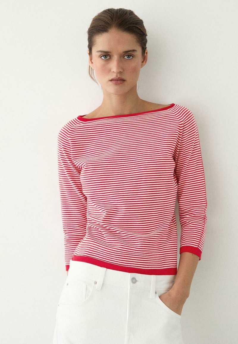 Kvinde med brunt hår iført en langærmet top med røde og hvide striber og hvide bukser, stående foran en ensfarvet, lys væg.