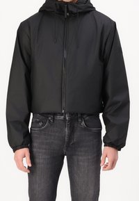 Schwarze cropped Jacke mit Kapuze, Frontreißverschluss, elastischen Bündchen und einer glatten, leichten Textur. Kombiniert mit dunklen Jeans.
