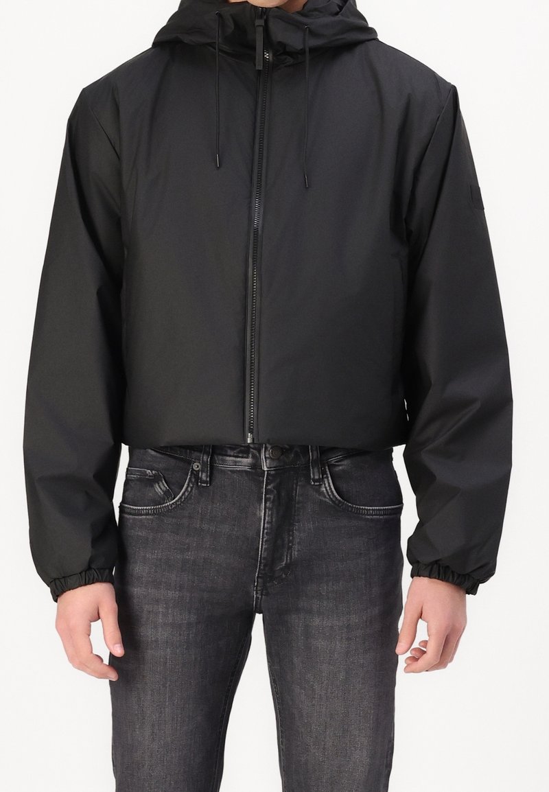 Schwarze cropped Jacke mit Kapuze, Frontreißverschluss, elastischen Bündchen und einer glatten, leichten Textur. Kombiniert mit dunklen Jeans.