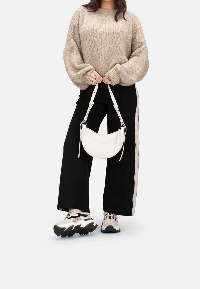 A person stands with their hands holding a small white handbag, wearing a cozy beige sweater and black wide-leg pants with side stripes.
