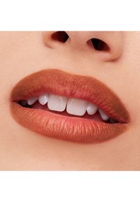 Matte leppestift i varm brun tone på glatte lepper. Viser full underleppe med definerte kanter og hvite tenner synlige. Nærbilde.