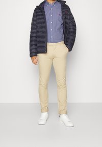 Námořnická bunda s podšívkou přes modrou kostkovanou košili, béžové slim-fit kalhoty a bílé tenisky. Bunda má zipové zapínání a kapucu.