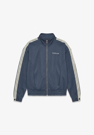 Tummansininen vetoketjullinen takki, jossa korkea kaulus, valkoiset virkatut raidat hihoissa, joustava vyötärönauha ja "PEGADOR"-logo vasemmassa rinnassa.