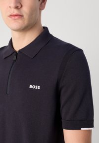 Σκούρο ναυτικό μπλουζάκι πόλο με κοντά μανίκια, υφή υφάσματος, μισό φερμουάρ και λευκό λογότυπο "BOSS" στο στήθος. Λεπτές ρίγες στους ώμους.