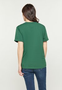 Grön bomullströja med uppvikt ärm, rund hals och mjuk textur. Bärs med mörkblå jeans och visar bakifrån.