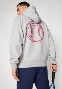 Grå hættetrøje med et grafisk design i pink cirkler på ryggen. Modellen holder en tennisketsjer og har mørke bukser på.