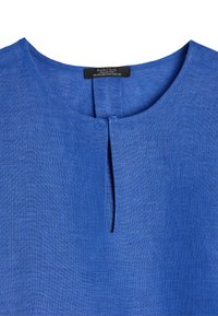 Niebieska lniana bluzka z okrągłym dekoltem, z przodem ozdobionym listwą i teksturowanym wykończeniem. Metka widoczna przy dekolcie.