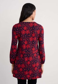 Raudonas gėlių raštas tunika su tamsiai mėlynu fonu. Alkūnių ilgio rankovės, apvalus iškirpimas ir surinkta tekstūra rankovių galuose.