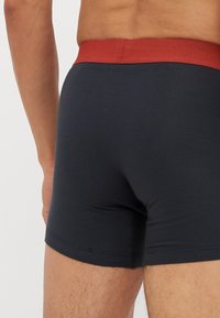 Shorts en boxeur de couleur bleu marine foncé avec une ceinture élastique rouge, présentant une texture lisse et un design ajusté, mettant en avant des ouvertures pour les jambes et des détails de couture.