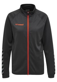 Veste noire zippée en tissu texturé, dotée d'une fermeture éclair orange et d'un logo. Agrémentée de rayures en chevron grises sur les manches.