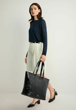 Kobieta w granatowej bluzce z długim rękawem, białe szorty z wysokim stanem, czarne buty na słupku, trzyma dużą czarną torebkę o fakturowanej powierzchni.