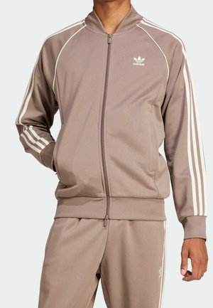 Mann trägt beigen Adidas-Trainingsanzug mit weißen Streifen an Ärmeln und Hosen, steht mit einer Hand in der Tasche und der anderen entspannt.